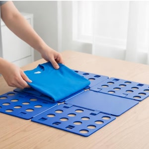 May include: A blue plastic garment folding board with a grid of circular cutouts, featuring a central solid panel and hinged side sections, currently being used to fold a bright blue t-shirt on a flat surface.