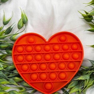 May include: Bright orange heart-shaped fidget toy with multiple circular bubbles. The toy is surrounded by green leafy sprigs on a white background. The bubbles are arranged in rows and columns.