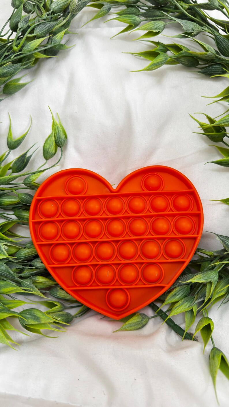 May include: Bright orange heart-shaped fidget toy with multiple circular bubbles. The toy is surrounded by green leafy sprigs on a white background. The bubbles are arranged in rows and columns.