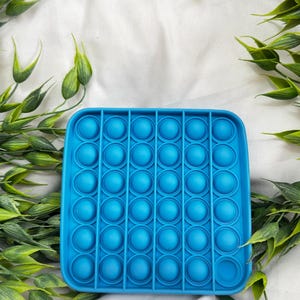 May include: A square, bright blue fidget toy with rounded edges and multiple circular bubble depressions. The toy is surrounded by green leafy sprigs, set against a white fabric background. This sensory toy is designed for stress relief.