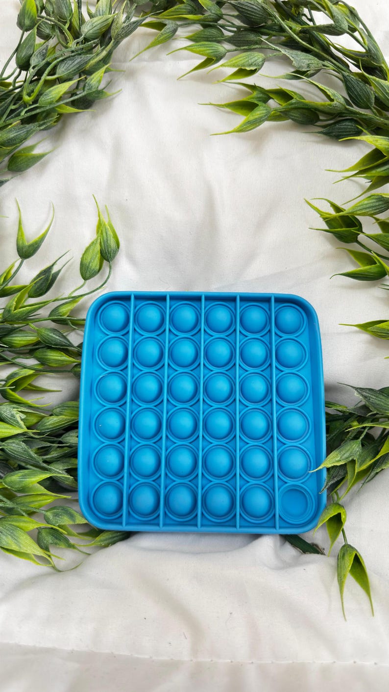 May include: A square, bright blue fidget toy with rounded edges and multiple circular bubble depressions. The toy is surrounded by green leafy sprigs, set against a white fabric background. This sensory toy is designed for stress relief.