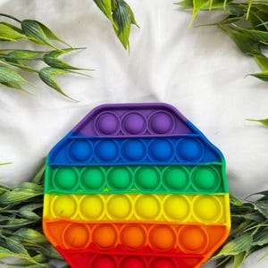 May include: A colorful, octagonal pop-it toy in a rainbow gradient of colors. The toy features rows of circular bubbles that can be pushed in and out. The background includes green leafy sprigs on a white surface.
