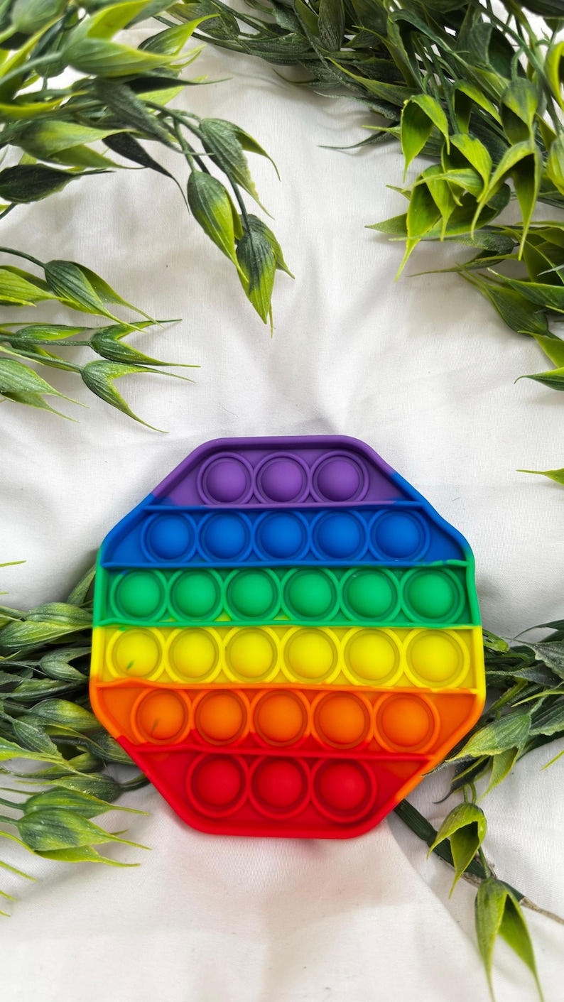 May include: A colorful, octagonal pop-it toy in a rainbow gradient of colors. The toy features rows of circular bubbles that can be pushed in and out. The background includes green leafy sprigs on a white surface.