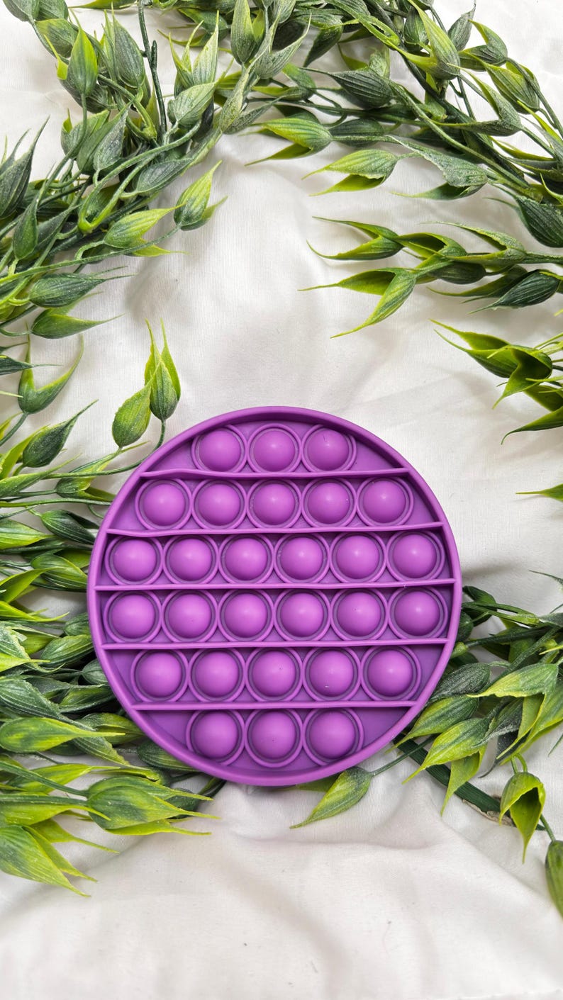 May include: A round, purple Pop It fidget toy with multiple bubble-like depressions. The toy is surrounded by green leafy sprigs against a white background. This sensory toy is designed for stress relief and tactile play.