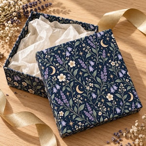 Könnte beinhalten: Eine quadratische Geschenkbox mit dunkelblauem Hintergrund und einem Muster aus violettem Lavendel, hellblauen glockenförmigen Blüten, weißen Blumen, goldenen Mondsicheln und kleinen Sternen. Die Box ist geöffnet und zeigt weißes Seidenpapier.