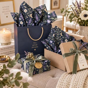 Könnte beinhalten: Eine dunkelblaue Geschenktüte mit goldenen Kordelgriffen und dem Text for good people, begleitet von zwei Geschenkboxen in dunklem Blumenpapier mit Monden und Sternen, verziert mit salbeigrünen Samtbändern und kleinen Trockenblumen.