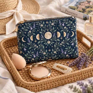 Puede incluir: Un estuche rectangular con cremallera hecho de tela azul oscuro con un patrón repetitivo de fases lunares, pequeñas flores blancas, ramas de lavanda y estrellas doradas. El estuche cuenta con un tirador de cremallera de metal dorado.