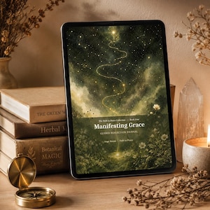 Könnte beinhalten: Ein digitales Tablet zeigt das Cover eines geführten Reflexionsjournals mit dem Titel "Manifesting Grace". Das Cover-Design zeigt ein himmlisches Design mit einem gewundenen goldenen Pfad. Ein Stapel Bücher, ein Kompass, eine Kerze und Trockenblumen sind auf einer Holzoberfläche angeordnet.