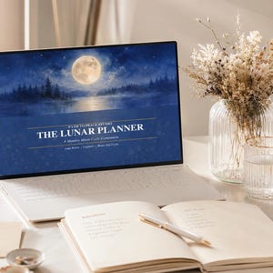 May include: A laptop screen displaying a digital planner titled THE LUNAR PLANNER by PATH TO PEACE STUDIO, featuring a full moon over a dark blue forest landscape. An open notebook with a pen rests in front of the laptop on a white surface.