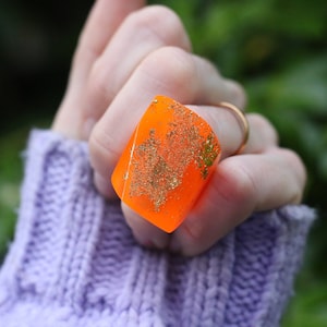 May include: A large, bright orange ring with gold glitter embedded in the resin. The ring is worn on a finger and is a statement piece of jewellery.