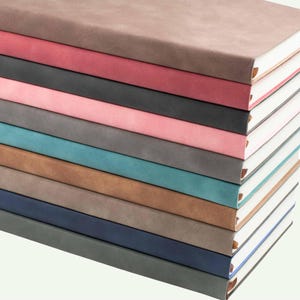 May include: A vertical stack of eight rectangular notebooks with matte, solid-coloured covers in shades of taupe, rose, charcoal, light pink, grey, teal, tan, and navy blue. Each notebook features white pages and a small, visible brown ribbon bookmark.