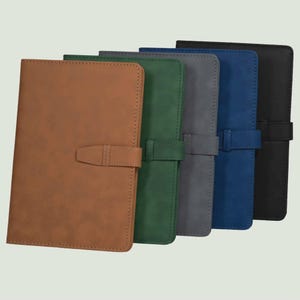 May include: A set of five rectangular notebooks with smooth covers in tan, dark green, gray, blue, and black. Each notebook features a matching horizontal strap closure with a stitched detail, arranged in a staggered, overlapping row.