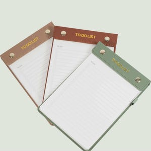 May include: Three rectangular to-do list notepads with white lined paper. Each notepad features a colored top header with two metallic studs and the text TO DO LIST printed in gold. The headers are light brown, dark brown, and sage green.