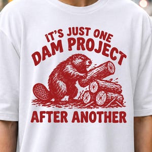 Könnte beinhalten: Ein weißes T-Shirt mit kurzen Ärmeln und einem roten Aufdruck eines Bibers, der Baumstämme stapelt. Der Text über dem Biber lautet IT'S JUST ONE DAM PROJECT und der Text darunter AFTER ANOTHER, alles in einer kräftigen, roten Schrift.