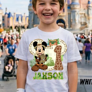 May include: White t-shirt with a Mickey Mouse safari design, the text "Birthday Boy", and the number "1". The name "Jaxson" is printed in green. The shirt is a birthday theme.