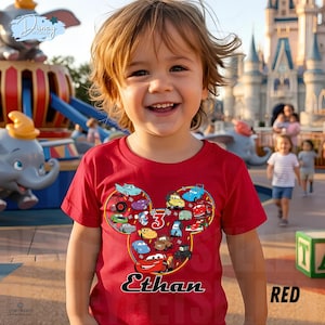 May include: A red t-shirt with a Mickey Mouse head design filled with cartoon cars. The shirt has the name "Ethan" in black script and the number "3". The background shows a theme park with a carousel and a castle.
