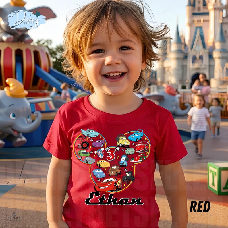 May include: A red t-shirt with a Mickey Mouse head design filled with cartoon cars. The shirt has the name "Ethan" in black script and the number "3". The background shows a theme park with a carousel and a castle.