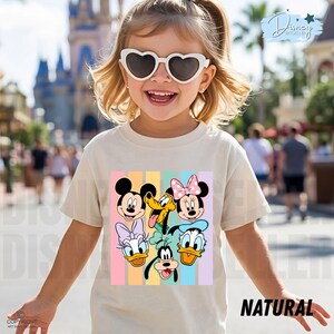 May include: A child wearing a cream-colored t-shirt featuring a colorful graphic of Disney characters, including Mickey Mouse, Minnie Mouse, Donald Duck, and Pluto. The word "NATURAL" is printed in black on the bottom right of the shirt.