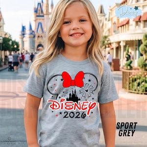 May include: A light gray t-shirt featuring a Minnie Mouse design with a red bow, fireworks, and a castle silhouette. The word "Disney" is in red, with "2026" below. The shirt is labeled "Sport Grey".