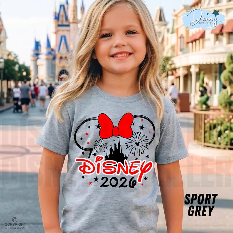 May include: A light gray t-shirt featuring a Minnie Mouse design with a red bow, fireworks, and a castle silhouette. The word "Disney" is in red, with "2026" below. The shirt is labeled "Sport Grey".