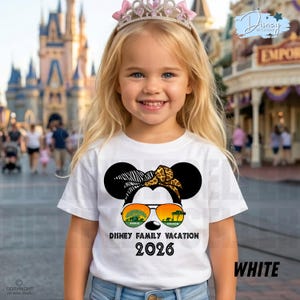 May include: A white t-shirt featuring a Minnie Mouse design with sunglasses and a headband. The shirt has the text "DISNEY FAMILY VACATION 2026" and "WHITE". A young girl is wearing the shirt and a pink tiara in front of a Disney park.