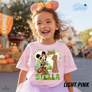 May include: A light pink t-shirt with a Minnie Mouse safari design, including the number 1, the name "Stella", and "Birthday Girl". The shirt is worn by a young girl with a headband featuring large ears. The text "LIGHT PINK" is visible.