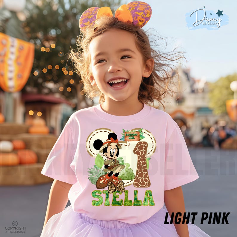 May include: A light pink t-shirt with a Minnie Mouse safari design, including the number 1, the name "Stella", and "Birthday Girl". The shirt is worn by a young girl with a headband featuring large ears. The text "LIGHT PINK" is visible.
