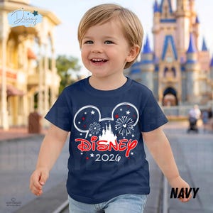 May include: A navy blue t-shirt featuring a Mickey Mouse head design with fireworks and a castle. The word "Disney" is in red script, with "2026" below. The shirt is worn by a smiling child.
