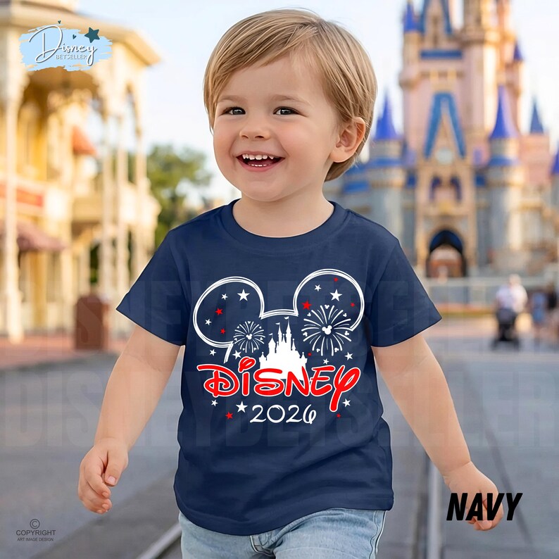 May include: A navy blue t-shirt featuring a Mickey Mouse head design with fireworks and a castle. The word "Disney" is in red script, with "2026" below. The shirt is worn by a smiling child.