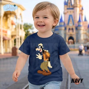 May include: A navy blue short-sleeved t-shirt featuring a graphic print of the cartoon character Goofy dressed in explorer attire, including a tan hat, orange vest, and khaki pants, with a backpack and brown shoes on a solid dark blue background.