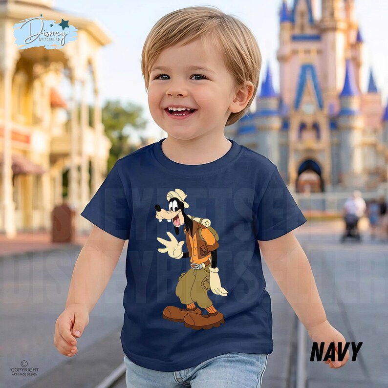 May include: A navy blue short-sleeved t-shirt featuring a graphic print of the cartoon character Goofy dressed in explorer attire, including a tan hat, orange vest, and khaki pants, with a backpack and brown shoes on a solid dark blue background.