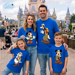 May include: A group of four people wearing matching royal blue t-shirts featuring Disney characters dressed in safari gear. The shirts display illustrations of Mickey Mouse, Goofy, Donald Duck, and Mickey Mouse again, all wearing hats and vests.