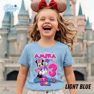 May include: A light blue t-shirt featuring a graphic of Minnie Mouse and Daisy Duck, the name Amira, and the number 3 in bright pink. The design includes colorful bunting and confetti accents, with the text Birthday Girl printed below the characters.