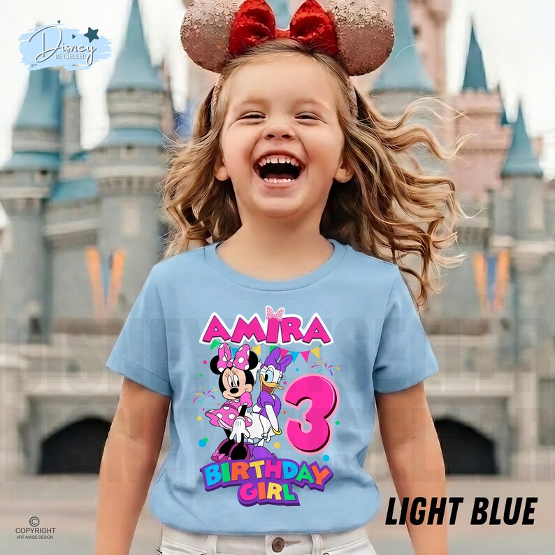 May include: A light blue t-shirt featuring a graphic of Minnie Mouse and Daisy Duck, the name Amira, and the number 3 in bright pink. The design includes colorful bunting and confetti accents, with the text Birthday Girl printed below the characters.