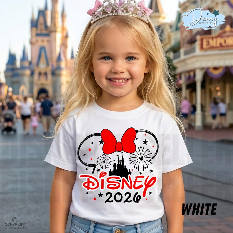 May include: A young girl wearing a white t-shirt with a Disney design and a pink tiara. The shirt features a red bow, Mickey Mouse ears, fireworks, a castle silhouette, and the word "Disney" and the year "2026" in red.