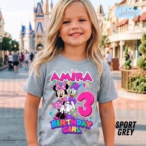 May include: A sport grey t-shirt featuring a graphic of Minnie Mouse and Daisy Duck, the name Amira, the number 3, and the text Birthday Girl. The design includes colorful confetti and bunting elements printed on the front of the garment.