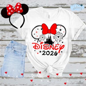 May include: White t-shirt with a Disney 2026 graphic featuring a red bow, fireworks, and a castle silhouette. A matching red bow headband with black mouse ears is next to the shirt, along with a pair of light blue jeans.