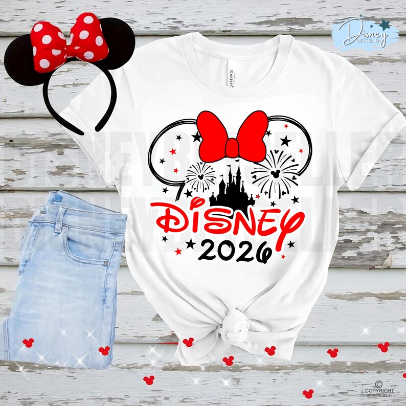 May include: White t-shirt with a Disney 2026 graphic featuring a red bow, fireworks, and a castle silhouette. A matching red bow headband with black mouse ears is next to the shirt, along with a pair of light blue jeans.
