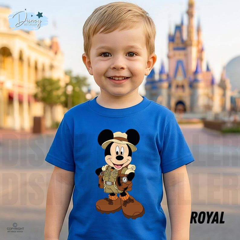 May include: A bright royal blue short-sleeved t-shirt featuring a graphic print of Mickey Mouse dressed in a tan safari outfit with a matching hat, brown boots, and a backpack. The shirt has a crew neckline and a straight hemline.