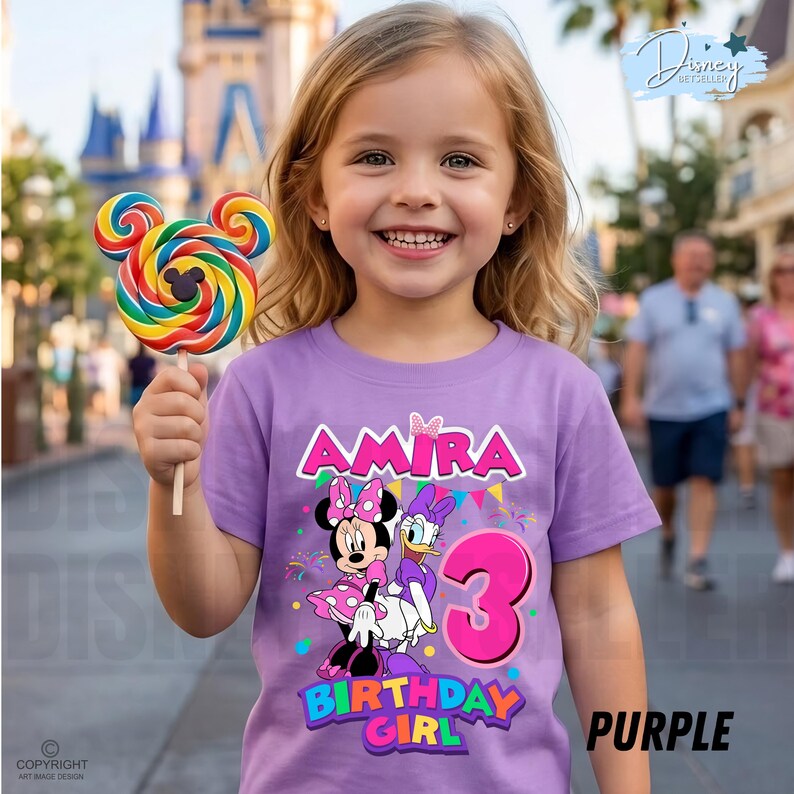 May include: A light purple t-shirt featuring a graphic of Minnie Mouse and Daisy Duck, the name Amira, and the number 3 in pink. The shirt includes colorful bunting and confetti designs. A multi-colored swirl lollipop is held in the foreground.