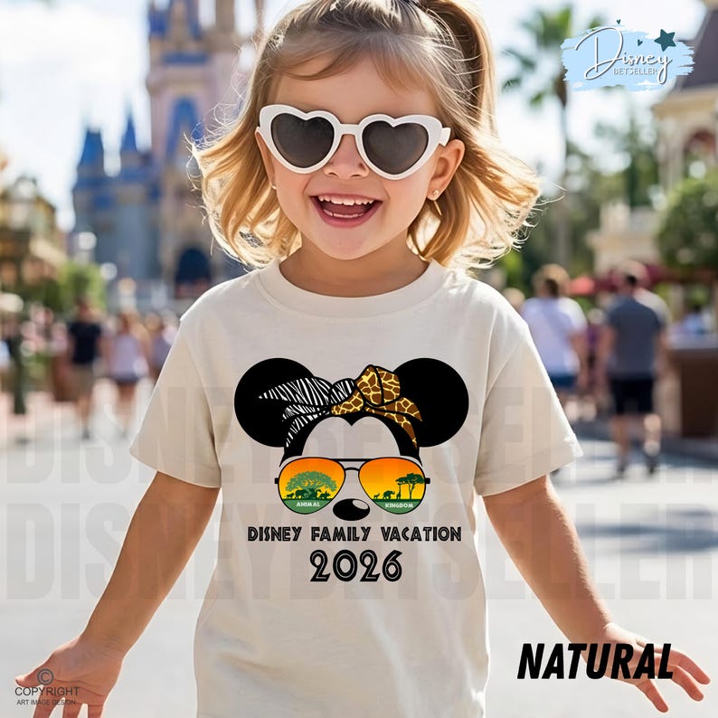 May include: A young girl wearing heart-shaped sunglasses and a beige t-shirt with a Minnie Mouse graphic. The graphic includes sunglasses with a safari scene, the text "DISNEY FAMILY VACATION 2026", and the word "NATURAL".