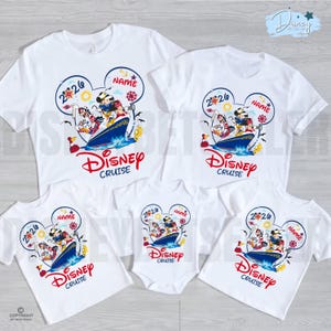 May include: A collection of white t-shirts and a baby bodysuit featuring a graphic of Disney characters on a cruise ship inside a Mickey Mouse ear outline. The design includes the text 2026, NAME, and Disney Cruise in red and blue colours.