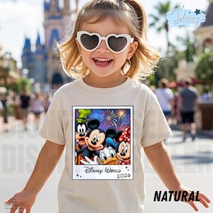 Puede incluir: Camiseta beige claro con un estampado colorido de Mickey, Minnie, Donald y Goofy, con fuegos artificiales y el texto "Disney World 2026". La camiseta est&aacute; etiquetada como "NATURAL". El ni&ntilde;o lleva gafas de sol en forma de coraz&oacute;n.
