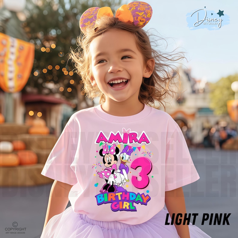 May include: A light pink t-shirt featuring a graphic of Minnie Mouse and Daisy Duck with the name Amira, the number 3, and the text Birthday Girl. The shirt is worn with a purple tulle skirt and a pair of orange and purple patterned mouse ears.