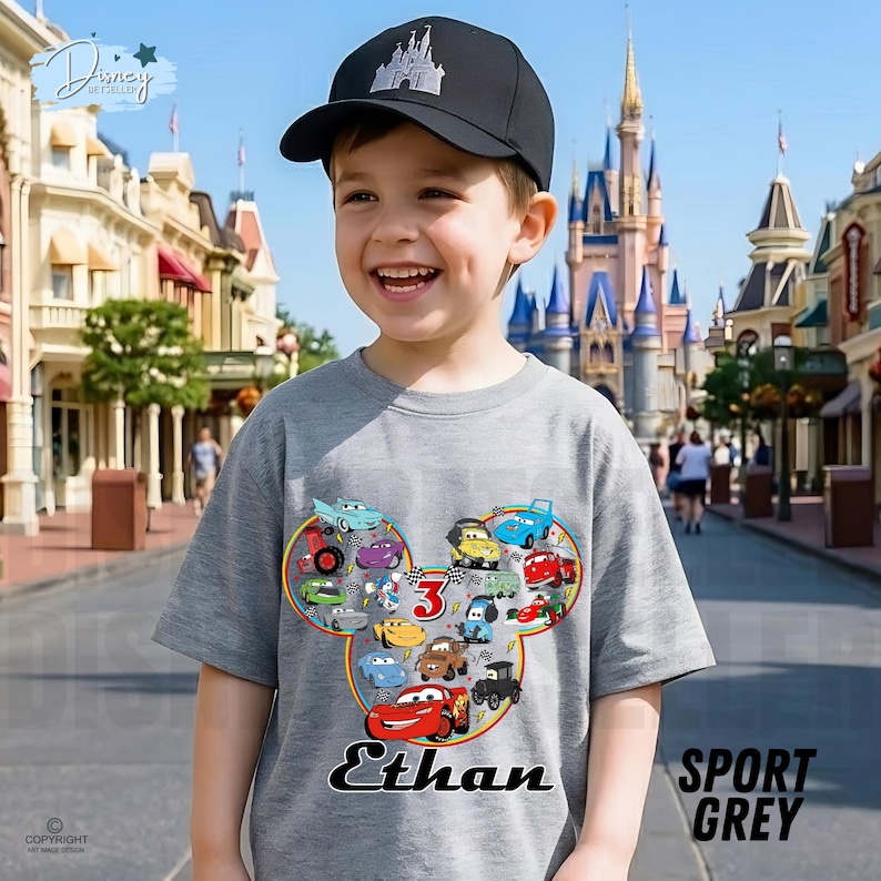 May include: A child in a grey t-shirt with a colourful cartoon car design in a Mickey Mouse head shape, with the name "Ethan" and the number "3". The child is also wearing a black baseball cap with a castle logo. The shirt is labelled "Sport Grey".