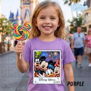 Puede incluir: Una persona joven sonr&iacute;e mientras sostiene una piruleta colorida con forma de Mickey Mouse. Lleva una camiseta morada con un gr&aacute;fico de Disney World 2026 con Mickey, Minnie, Donald y Goofy. La palabra "PURPLE" est&aacute; impresa en la camiseta.