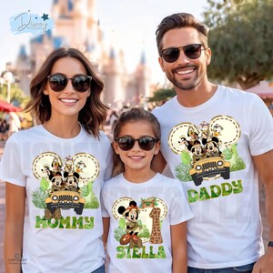 May include: White t-shirts with a Mickey Mouse safari design. The shirts feature Mickey, Donald, Goofy, and Pluto in a jeep. One shirt says "Mommy", another says "Daddy", and the third says "Stella" with a number 1 and a giraffe.
