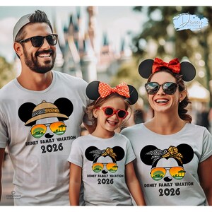 May include: Three people wearing matching light gray t-shirts with a Mickey Mouse graphic. The graphic includes sunglasses, a safari hat, and the text "DISNEY FAMILY VACATION 2026". The family is wearing Mickey Mouse ears and sunglasses.