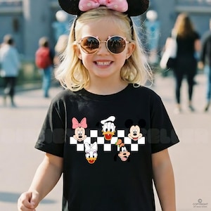 以下が含まれることがあります： 黒いTシャツ。市松模様のデザインで、ミニーマウス、ドナルドダック、ミッキーマウス、デイジーダック、グーフィーなどの漫画のキャラクターの顔が描かれています。サングラスとミッキーマウスの耳をつけた人が着ています。