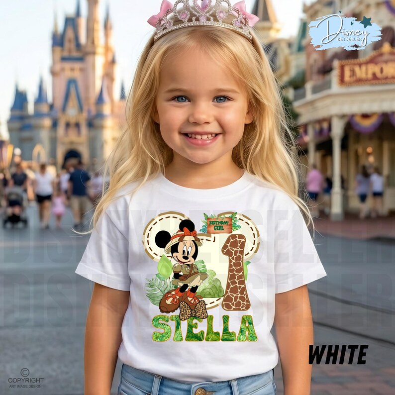 May include: A young girl wearing a white t-shirt featuring a Minnie Mouse design, a pink tiara, and the name "Stella". The shirt design includes a giraffe-patterned number 1 and the words "Birthday Girl".